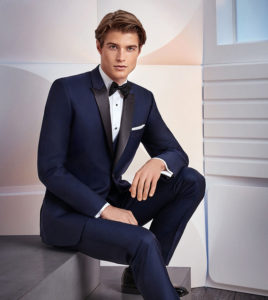 Here's an 8-word alt tag for the image: Man in navy tuxedo, sitting, black bow tie.